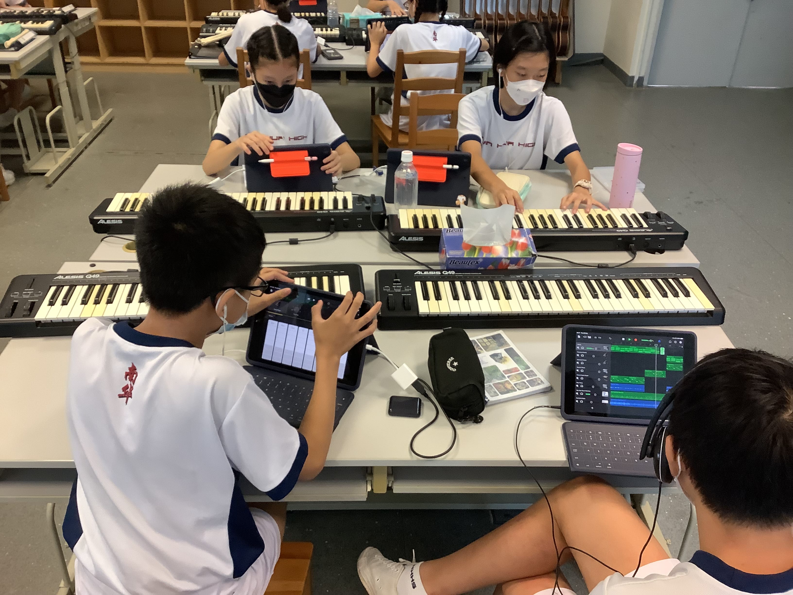 Students in white uniforms use Alesis MIDI keyboards and tablets in a music classroom.